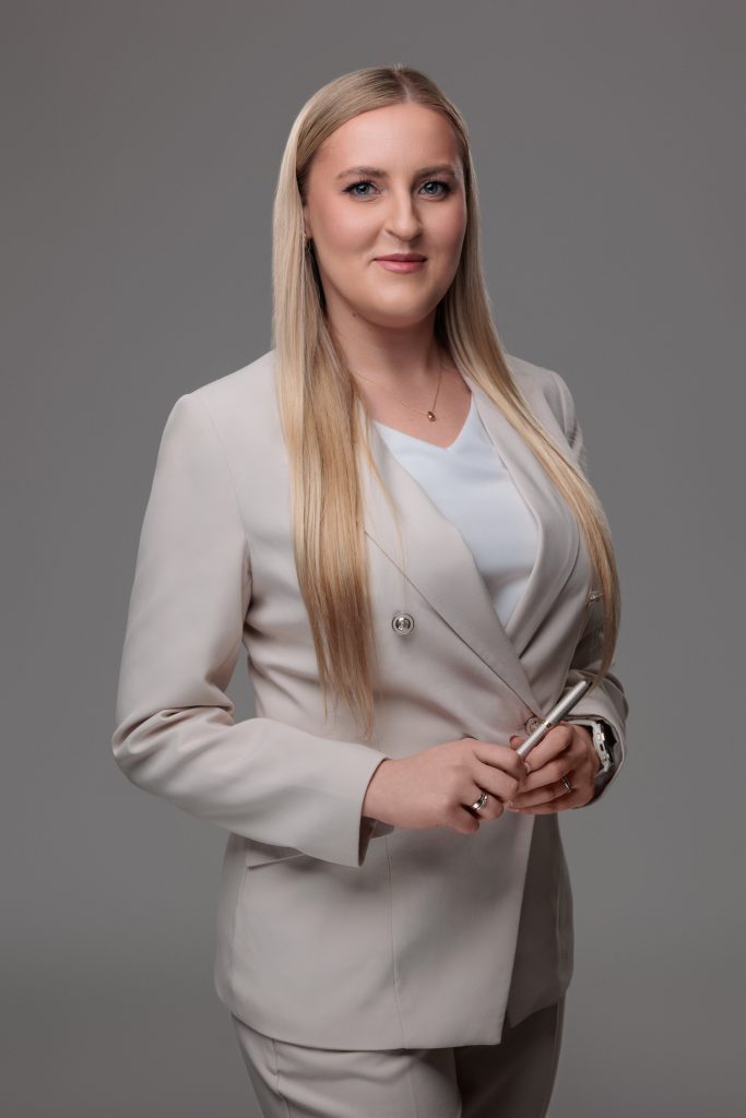 Zdjęcie biznesowe uśmiechniętej blondynki w beżowym garniturze i białej bluzce. Kobieta stoi na szarym tle, trzymając w dłoniach eleganckie pióro.