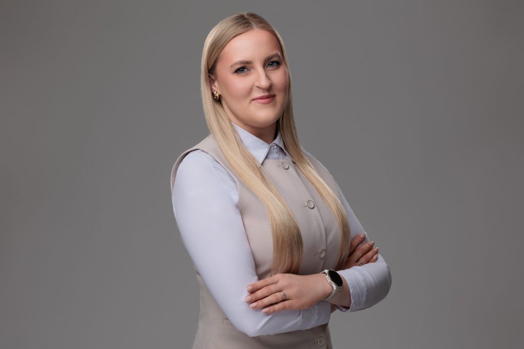 Portret uśmiechniętej kobiety o blond włosach, ubranej w białą koszulę i beżową kamizelkę, stojącej z założonymi rękami na szarym tle.